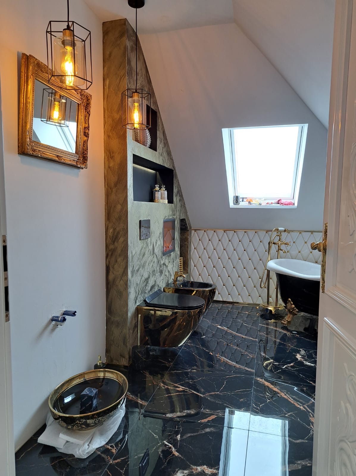 Luksusowa łazienka na poddaszu z czarno-złotymi płytkami, wanną na lwich nóżkach i złotymi akcentami, oświetlona wiszącymi lampami w stylu industrialnym.
