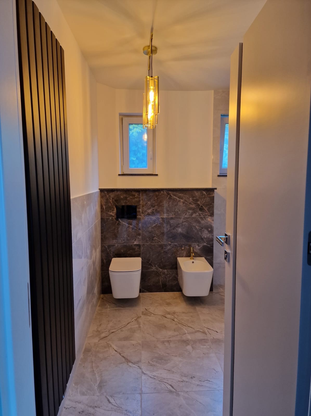 Nowoczesna łazienka z marmurową ścianą i podłogą w odcieniach szarości. Wisząca toaleta i bidet, designerska lampa. Minimalistyczny grzejnik żeberkowy.