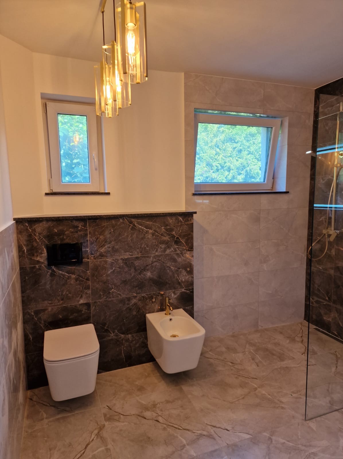 Wykończona łazienka z marmurowymi płytkami, wiszącą toaletą i bidetem. Nad nimi okno i ozdobna lampa. Widoczny fragment szklanej kabiny prysznicowej.