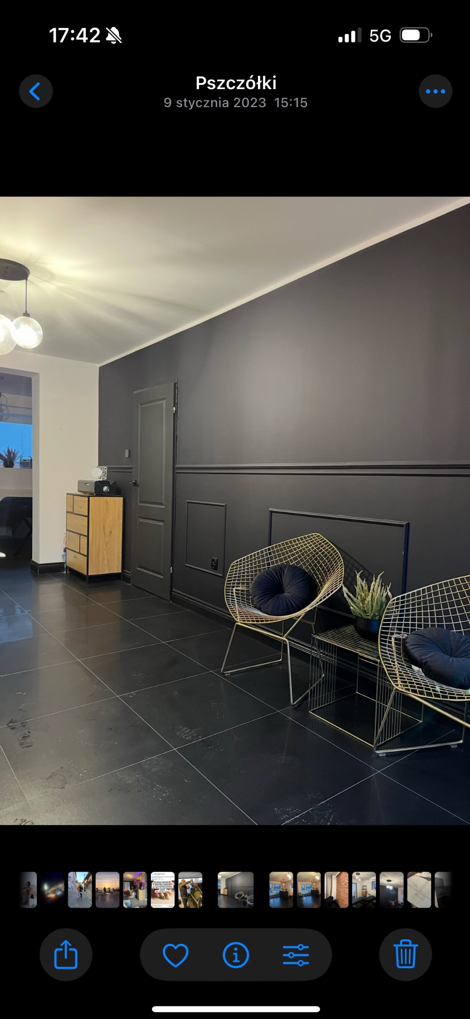 Czarne płytki podłogowe w salonie, ściana z lameli w kolorze grafitowym, złote krzesła z poduszkami, drewniana komoda i drzwi w odcieniach szarości.