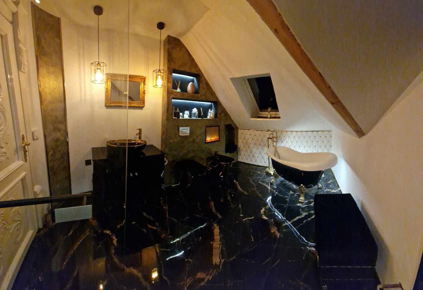 Luksusowa łazienka na poddaszu z czarną marmurową podłogą, wolnostojącą wanną na złotych nóżkach i umywalką na czarnej szafce. Oświetlenie punktowe i dekoracyjne nisze.