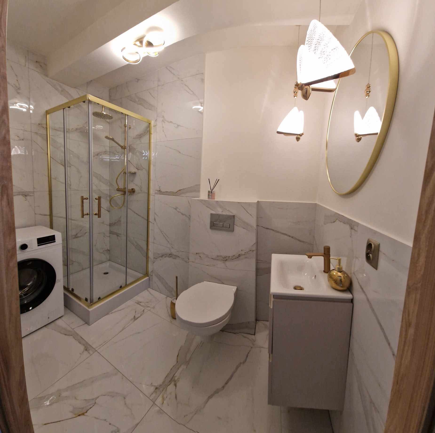 Elegancka łazienka w bieli z akcentami złota: kabina prysznicowa, umywalka z szafką, toaleta i pralka. Marmurowe płytki podłogowe i ścienne. Designerskie oświetlenie.