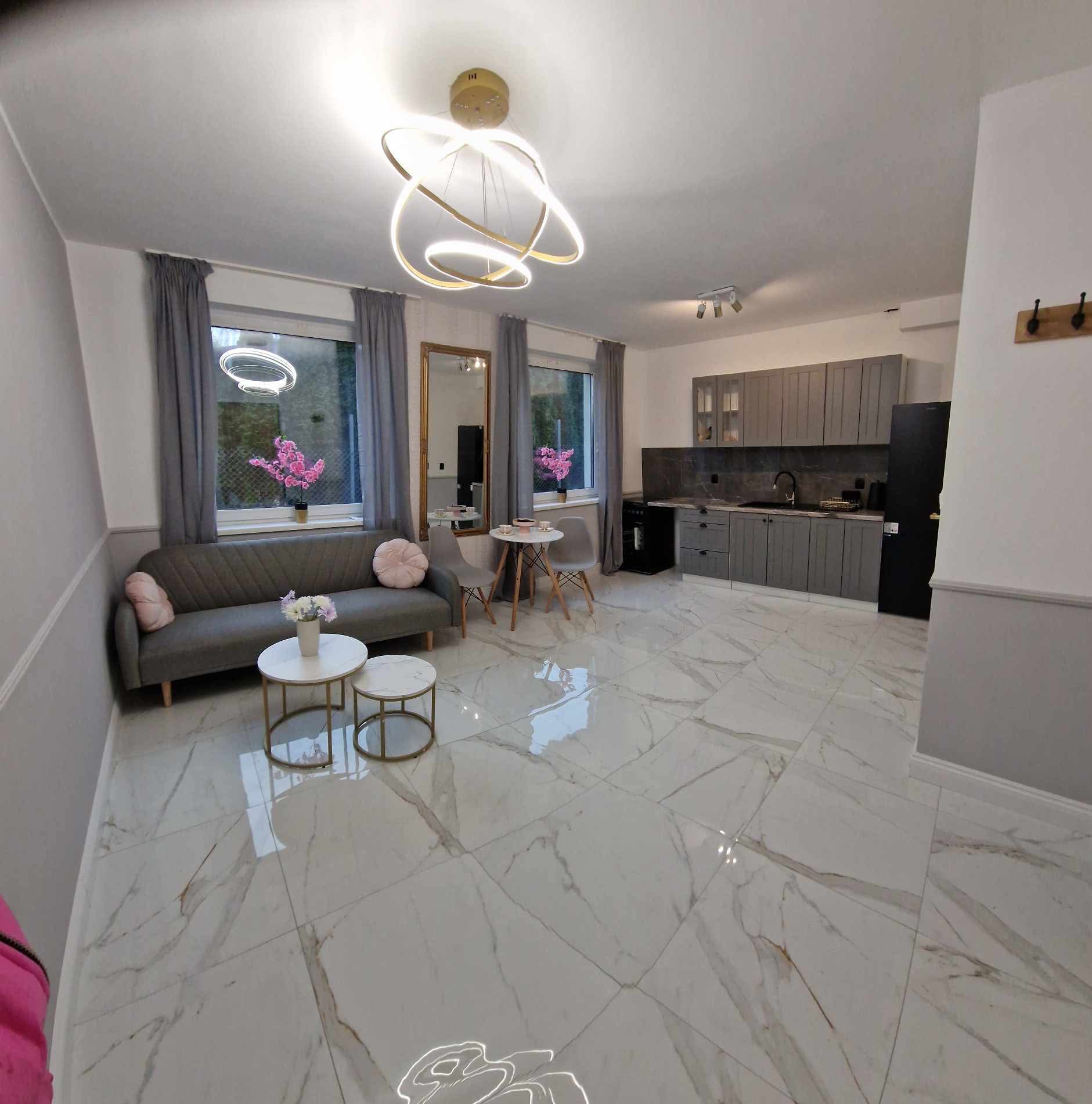 Jasne, przestronne wnętrze z białymi marmurowymi płytkami podłogowymi. Szara sofa, okna z zasłonami i kuchnia w tle. Nowoczesna lampa sufitowa.