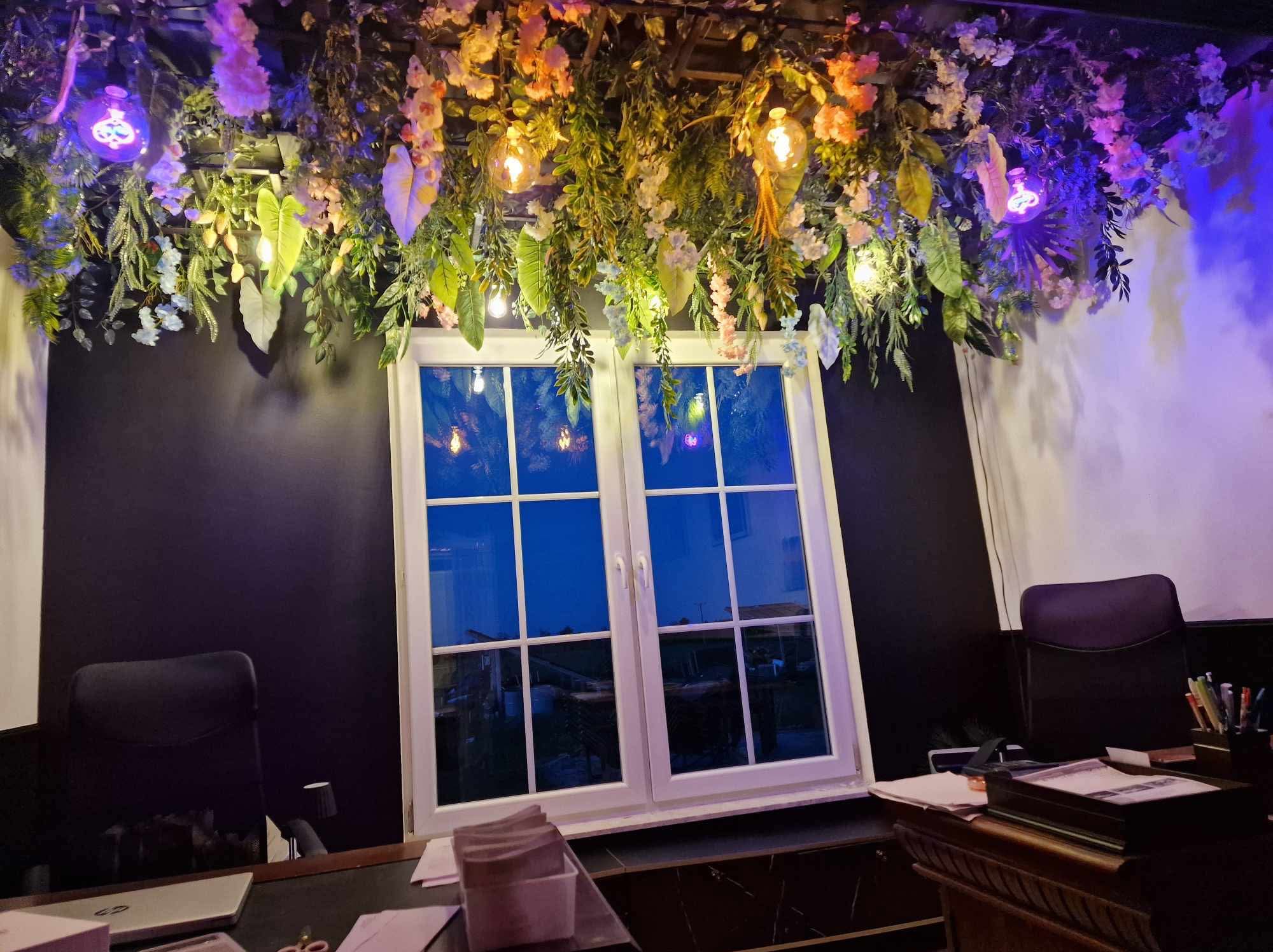 Ciemne biuro z oknem, dekoracją kwiatową i lampami. Widoczne biurko i krzesła. Nietypowe oświetlenie i wystrój wnętrza.