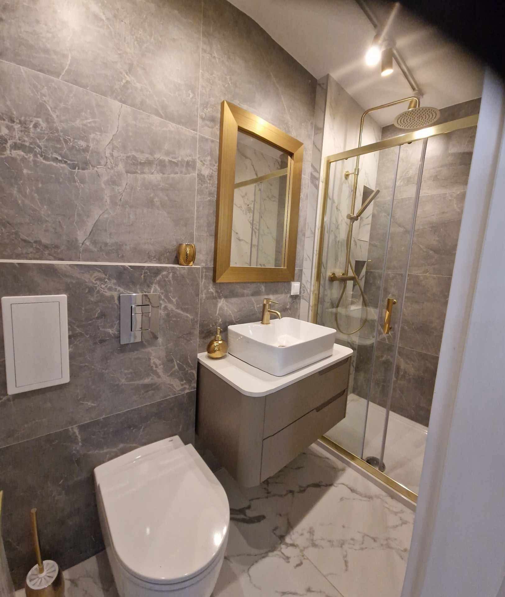 Elegancka łazienka z szarymi marmurowymi płytkami, złotymi akcentami i nowoczesną armaturą. Umywalka nablatowa, kabina prysznicowa typu walk-in.