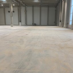 Poler-floor Mateusz Czarnecki - Surowa, betonowa posadzka przemysłowa w hali magazynowej, widoczne ślady po maszynach. W tle dwóch pracowników. Duże okno z prawej strony.