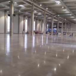 Poler-floor Mateusz Czarnecki - Błyszcząca, szara posadzka przemysłowa w przestronnej hali z oknami i betonowymi kolumnami. Widoczny sprzęt do polerowania i pracownicy w oddali.