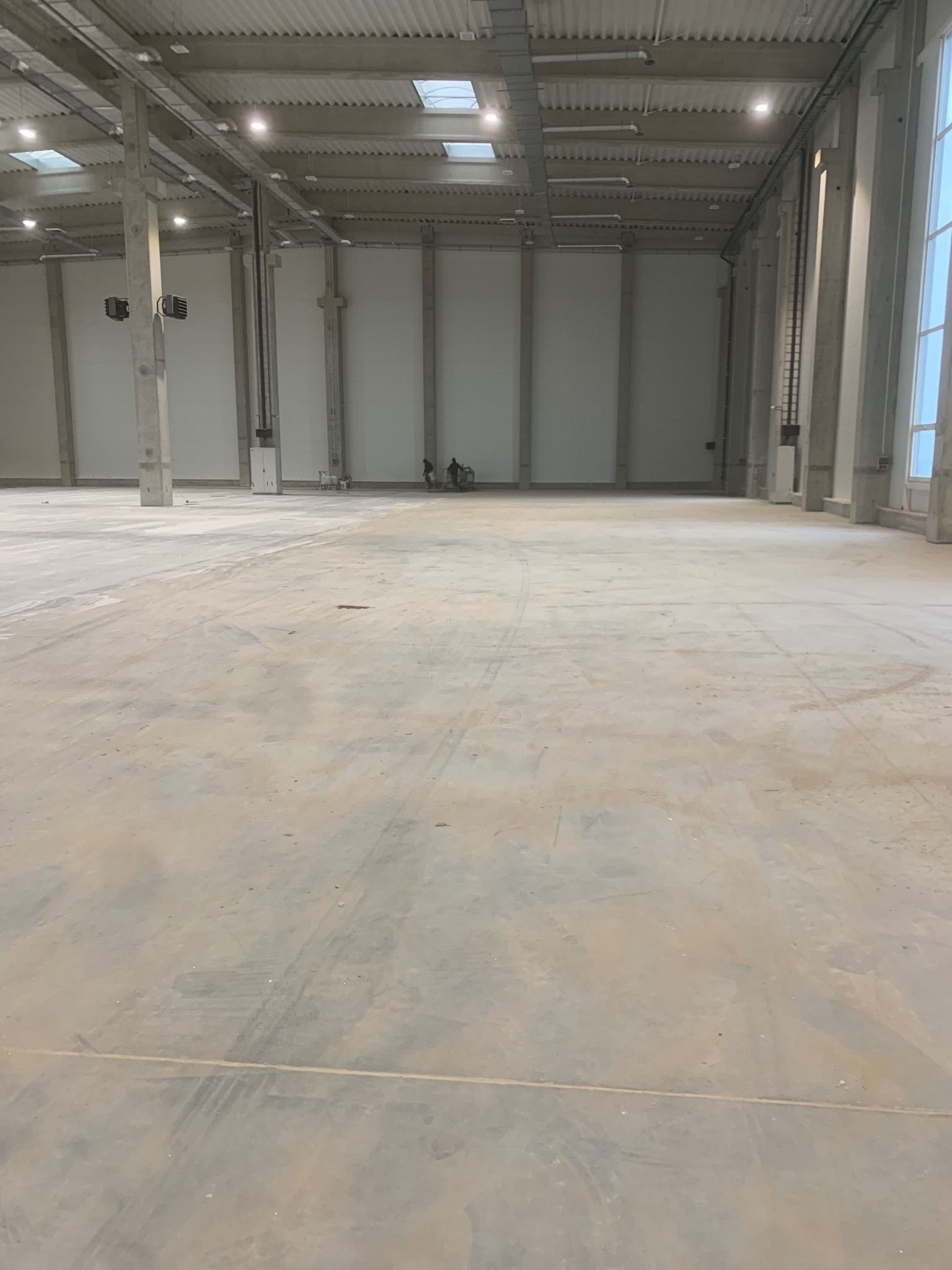 Surowa, betonowa posadzka przemysłowa w hali magazynowej, widoczne ślady po maszynach. W tle dwóch pracowników. Duże okno z prawej strony.