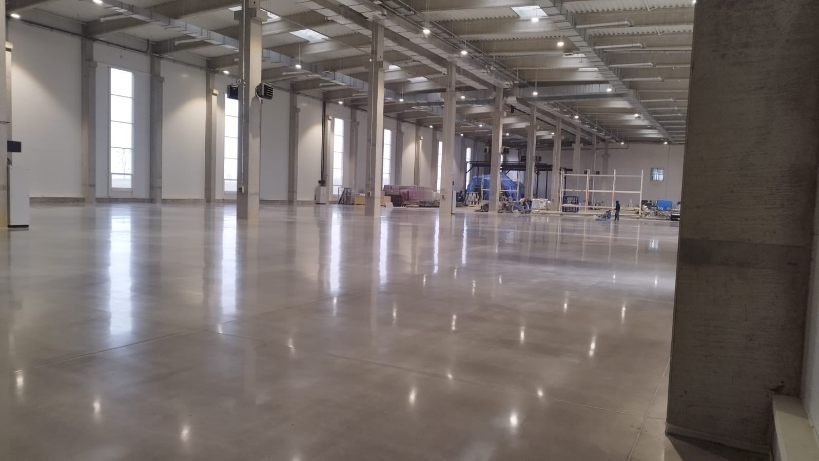 Błyszcząca, szara posadzka przemysłowa w przestronnej hali z oknami i betonowymi kolumnami. Widoczny sprzęt do polerowania i pracownicy w oddali.