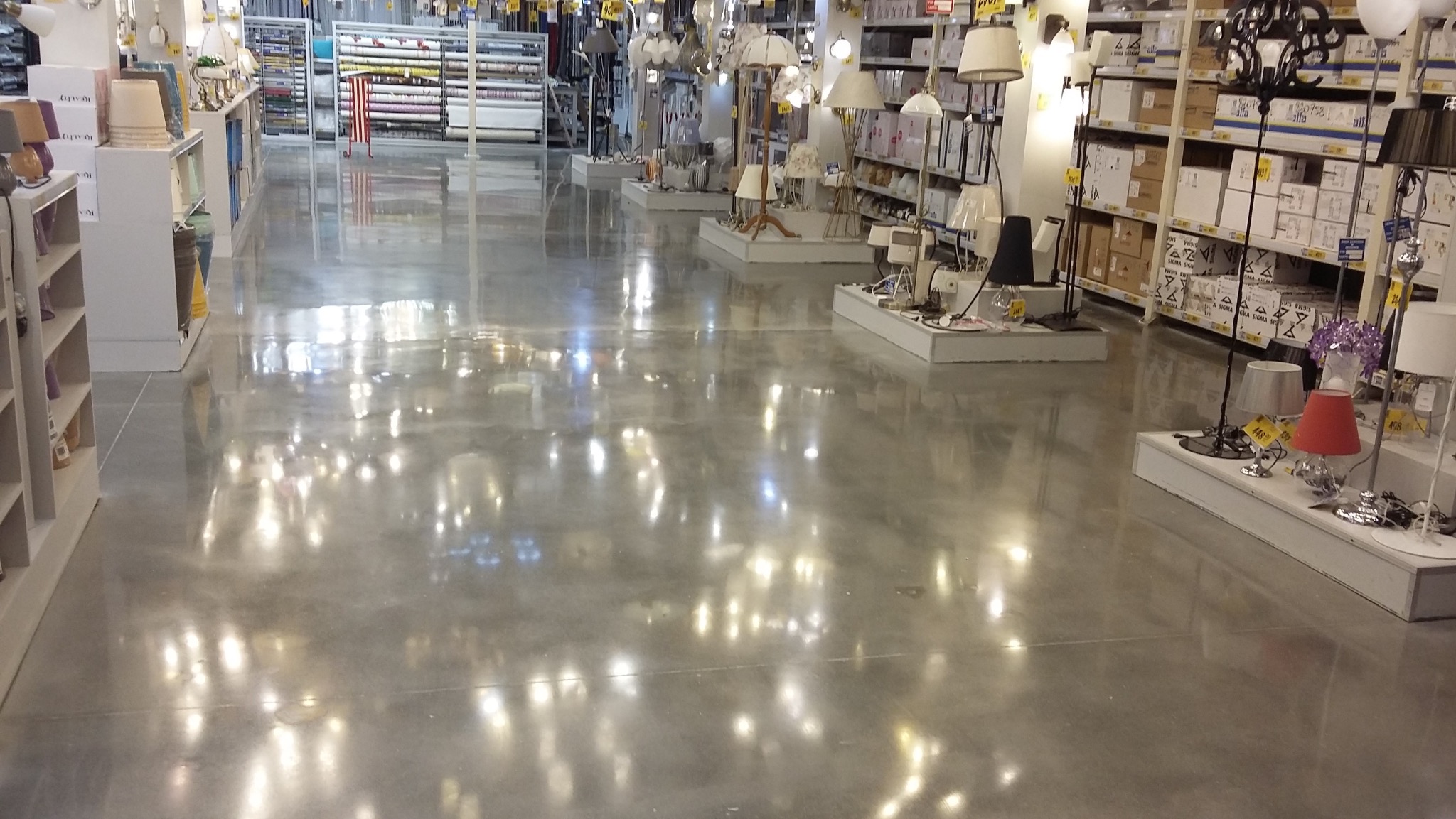 Błyszcząca, szara posadzka przemysłowa w sklepie oświetleniowym, odbijająca światła lamp. Widoczne regały z lampami i elementami wyposażenia sklepu.