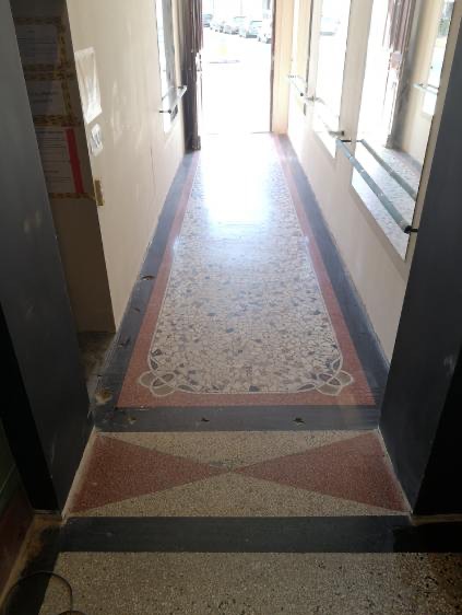 Korytarz z posadzką lastryko w odcieniach szarości, bieli i czerwieni. Widoczne ślady użytkowania i zabrudzenia. W tle otwarte drzwi.