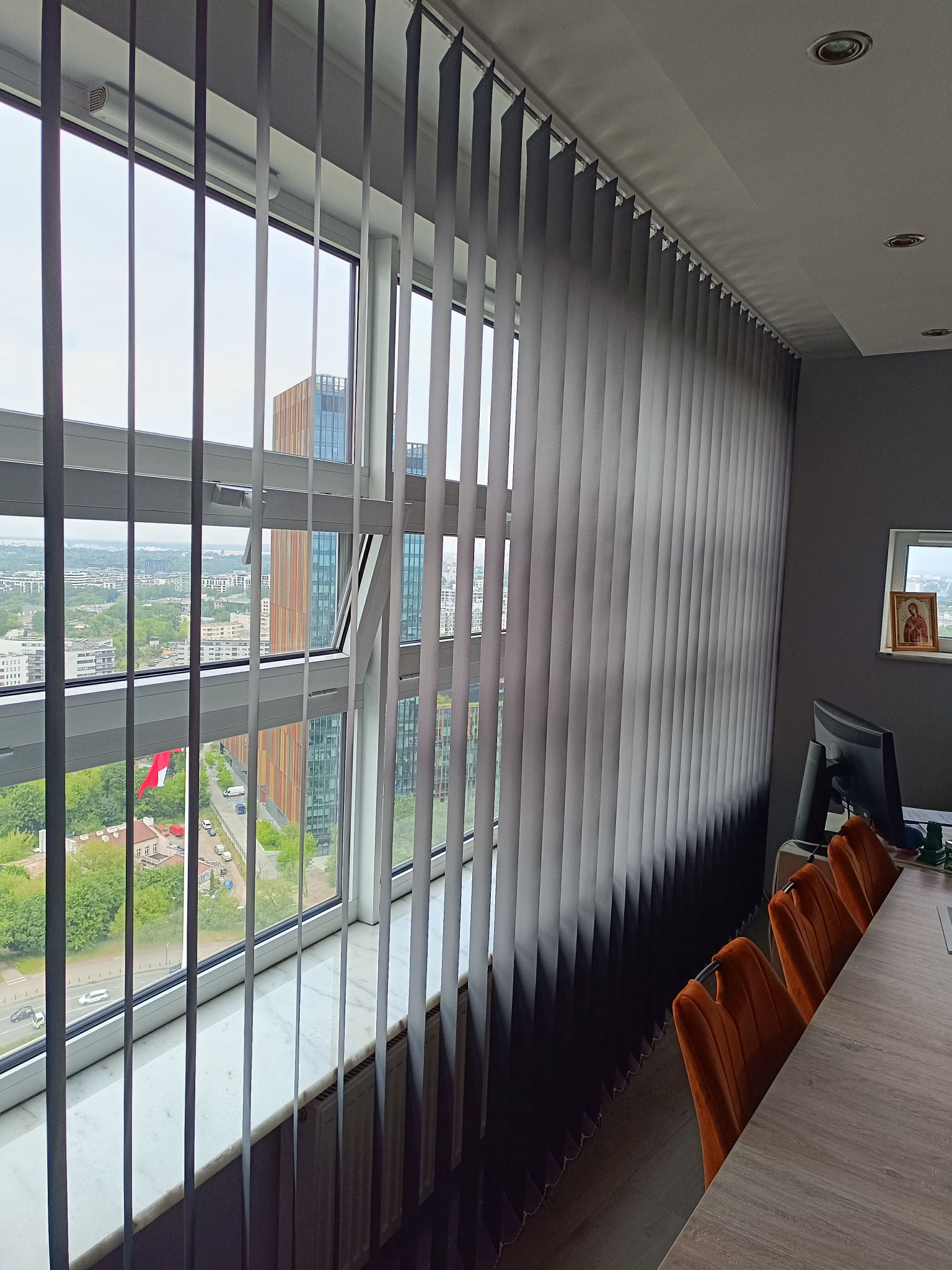 Szare żaluzje pionowe w biurze, widok na miasto przez okno. Częściowo widoczne pomarańczowe krzesła i biurko. Na ścianie ikona w ramce.