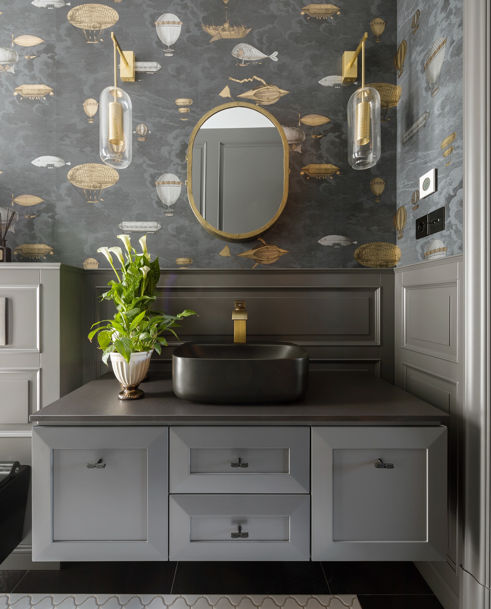 Stylowa łazienka z szarą szafką pod umywalkę, czarną umywalką nablatową, złotą armaturą i lustrem w złotej ramie. Ściany zdobi tapeta z motywem balonów.