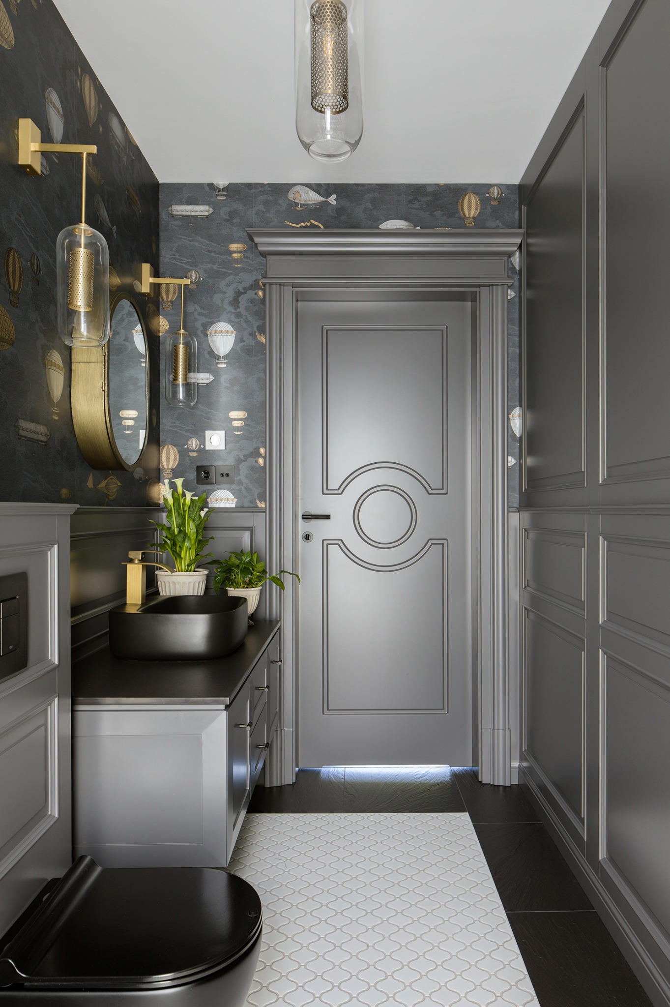 Stylowa, szara łazienka z czarną umywalką nablatową i złotą armaturą. W tle tapeta w balony i ryby, okrągłe lustro i designerskie lampy. Uwagę przykuwa wzór podłogi.