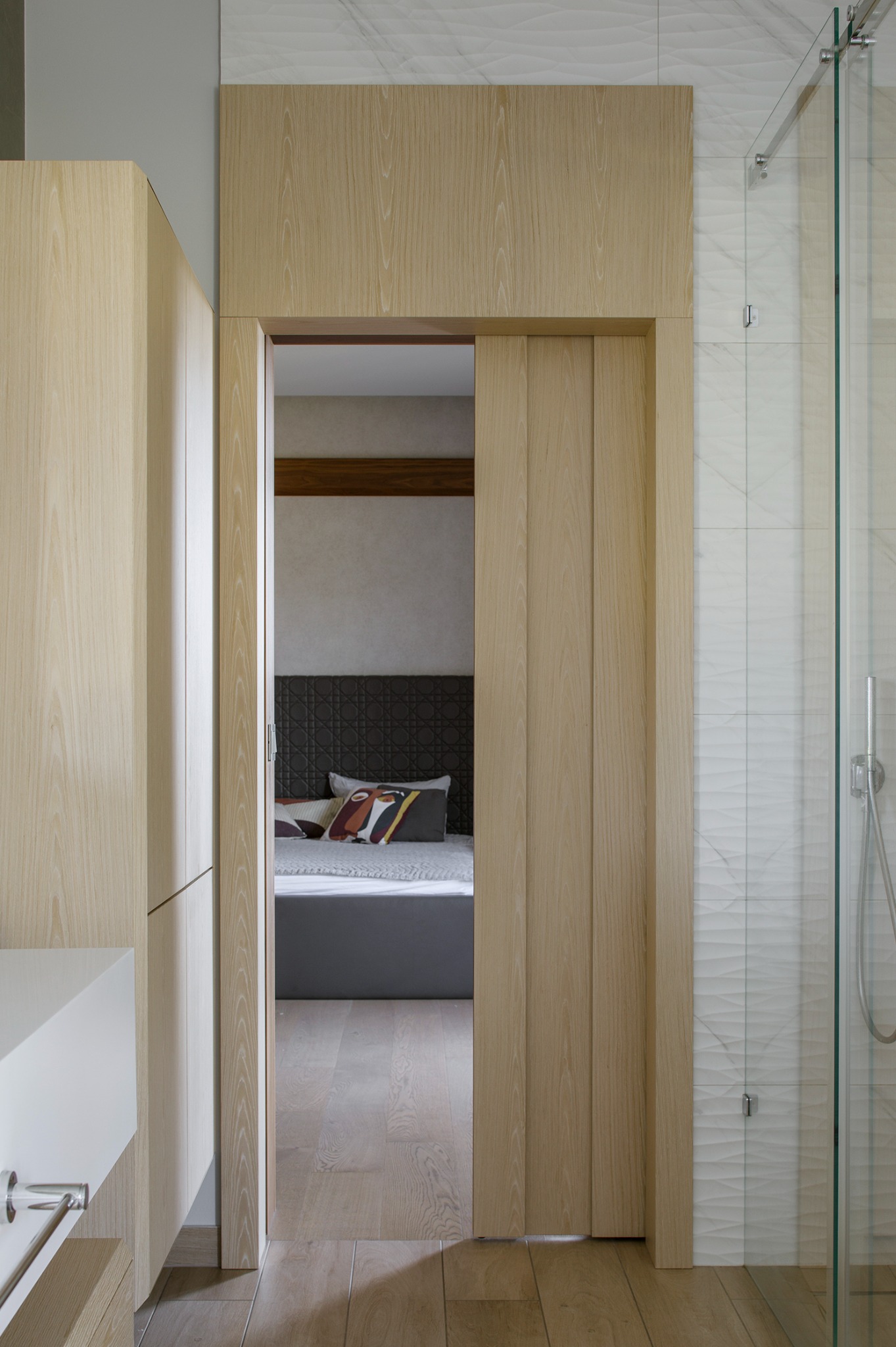 Nowoczesna łazienka z drewnianymi elementami: przesuwna zabudowa, płytki imitujące marmur i widok na sypialnię z łóżkiem i poduszkami dekoracyjnymi.