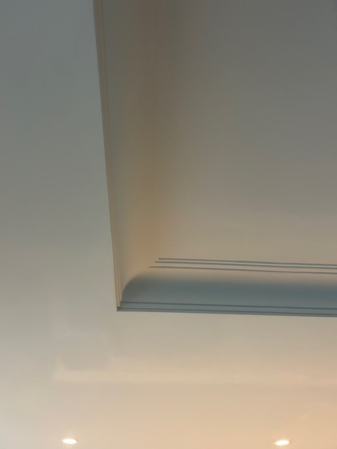 Biały sufit z dekoracyjnym, profilowanym gzymsem w rogu pomieszczenia. Widoczne dwa punkty oświetleniowe w tle. Minimalistyczny design.