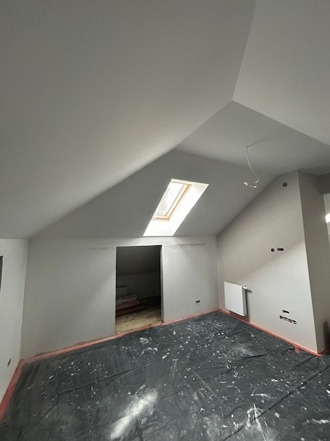 Poddasze po gładziowaniu ścian z oknem dachowym i zabezpieczoną folią podłogą. Widoczne przygotowania do dalszych prac wykończeniowych.