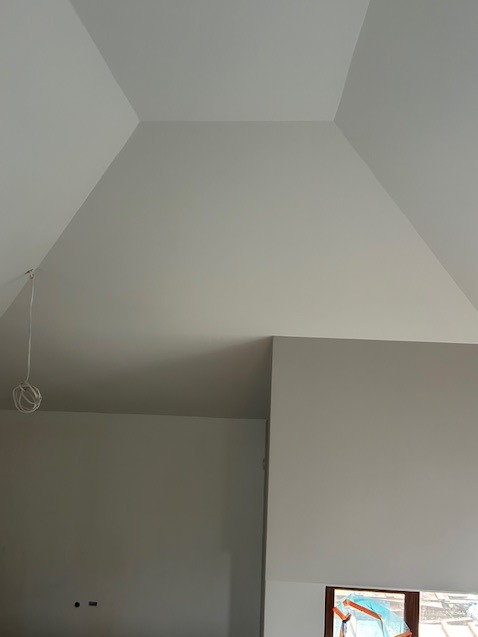 Wysoki sufit ze skosami, świeżo pomalowany na biało. Widoczne gniazdka elektryczne i wystające kable. Okno zabezpieczone folią malarską.