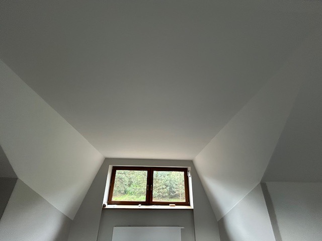 Poddasze po remoncie w Leoncinie: świeżo pomalowane ściany i sufit, widoczne okno z widokiem na zieleń. Skośne ściany pomalowane na biało.