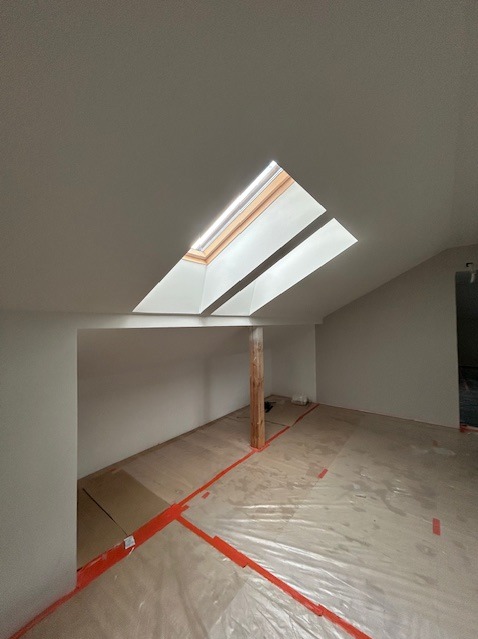 Poddasze po remoncie z oknem dachowym i zabezpieczoną podłogą folią malarską. Widoczny słup podtrzymujący strop. Ściany pomalowane na biało.