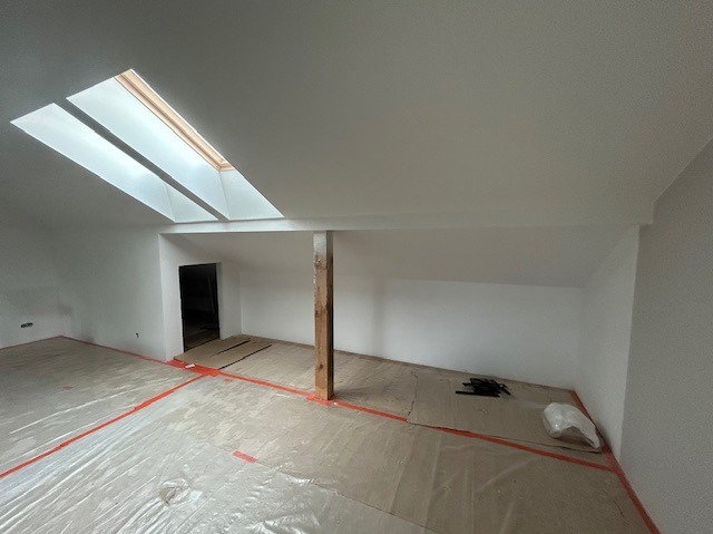 Białe poddasze po remoncie, zabezpieczone folią malarską, z drewnianą kolumną i oknami dachowymi. Widok na puste, odnowione wnętrze przed dalszym wykończeniem.