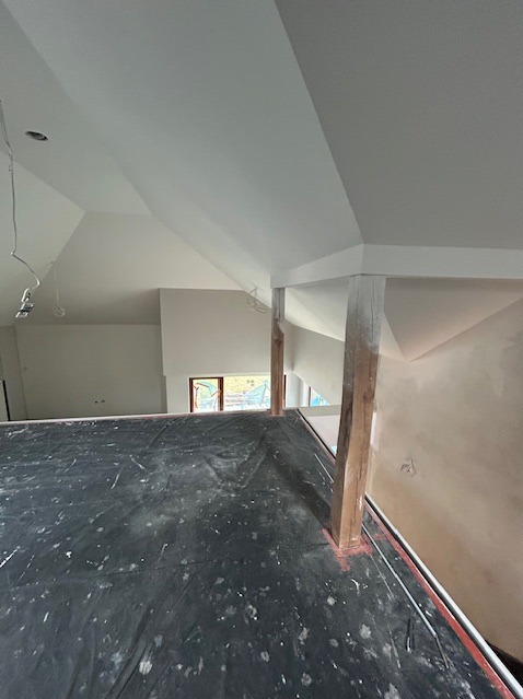 Wnętrze w trakcie malowania: białe ściany, drewniane słupy. Podłoga zabezpieczona czarną folią malarską. Widoczne okno i kable wystające ze ściany.