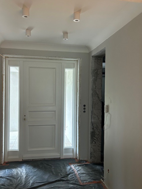 Białe, dwuskrzydłowe drzwi wejściowe z przeszkleniami bocznymi w trakcie montażu, zabezpieczone folią. Widoczne oświetlenie sufitowe i fragment ściany.