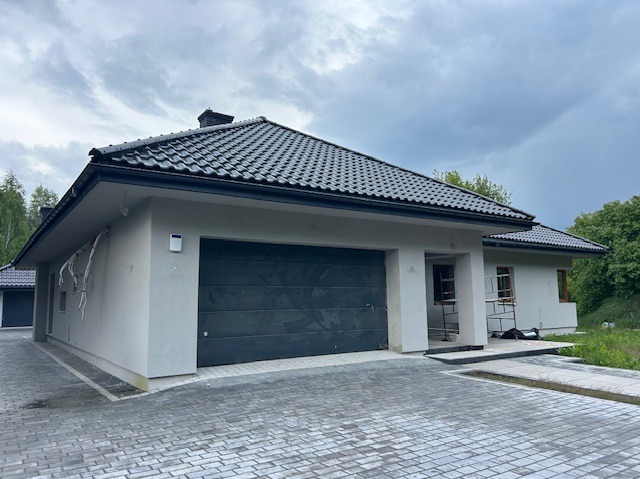 Parterowy dom z grafitową bramą garażową i kostką brukową na podjeździe. Częściowo otynkowana elewacja i czarna dachówka. Widoczne rusztowania przy oknach.