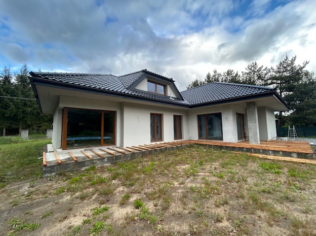 Nowoczesny dom jednorodzinny z ciemną dachówką i dużymi oknami. Widoczne przygotowania do budowy tarasu. Otoczenie: trawa, drzewa, pochmurne niebo.