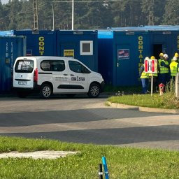 DOK-STER LP ODDZIAŁ W POLSCE - Biały samochód dostawczy z logo Dom-Ster na tle niebieskich kontenerów biurowych i grupy osób w kaskach i kamizelkach odblaskowych. Kontenery z napisami.