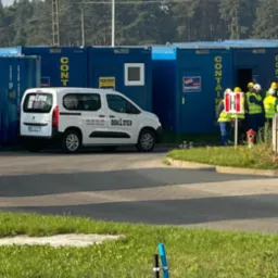 Biały samochód dostawczy z logo Dom-Ster na tle niebieskich kontenerów biurowych i grupy osób w kaskach i kamizelkach odblaskowych. Kontenery z napisami.