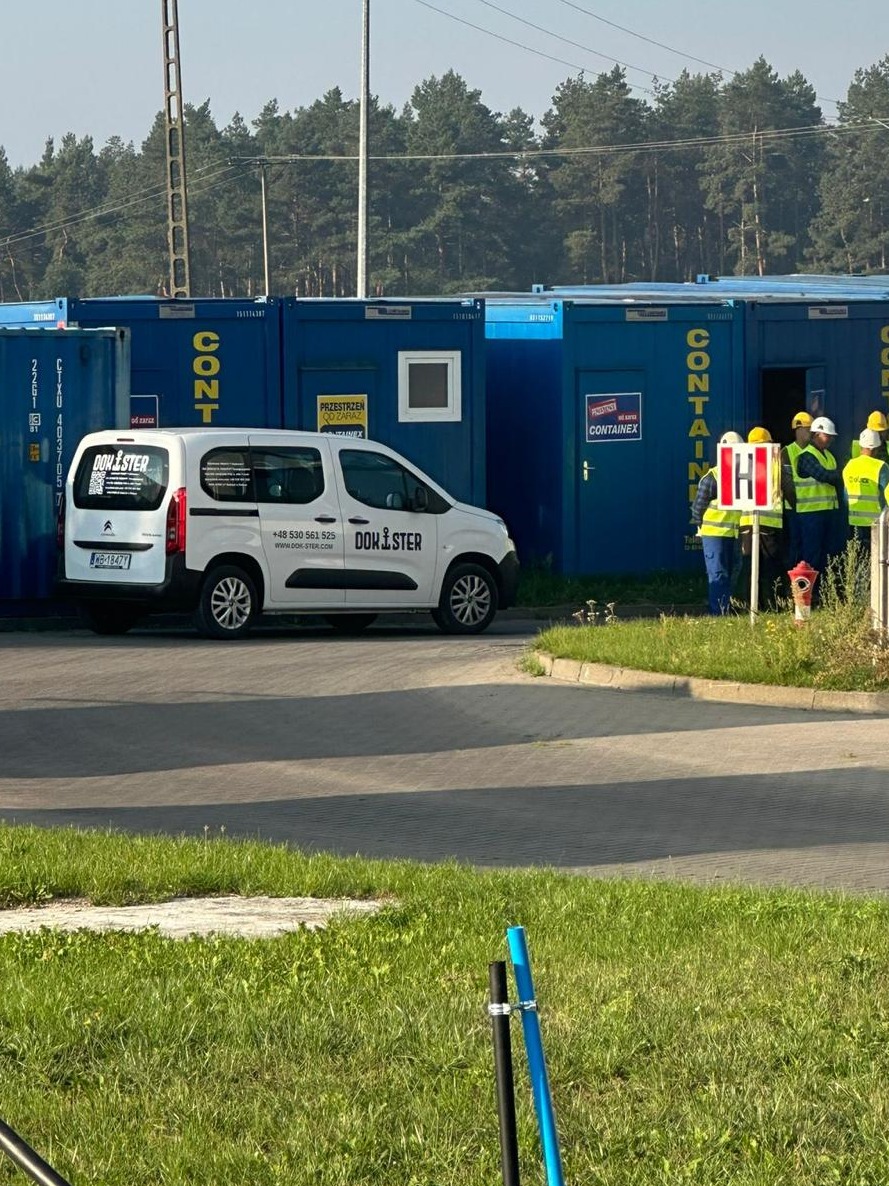 Biały samochód dostawczy z logo Dom-Ster na tle niebieskich kontenerów biurowych i grupy osób w kaskach i kamizelkach odblaskowych. Kontenery z napisami.