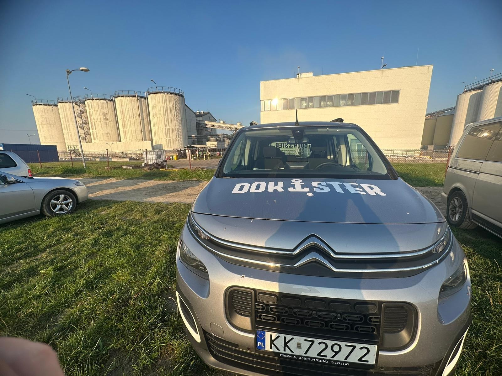 Szary Citroen Berlingo z napisem 'DOKISTER' na masce, zaparkowany na trawie, w tle silosy i budynki przemysłowe w słoneczny dzień. Tablica rejestracyjna widoczna.