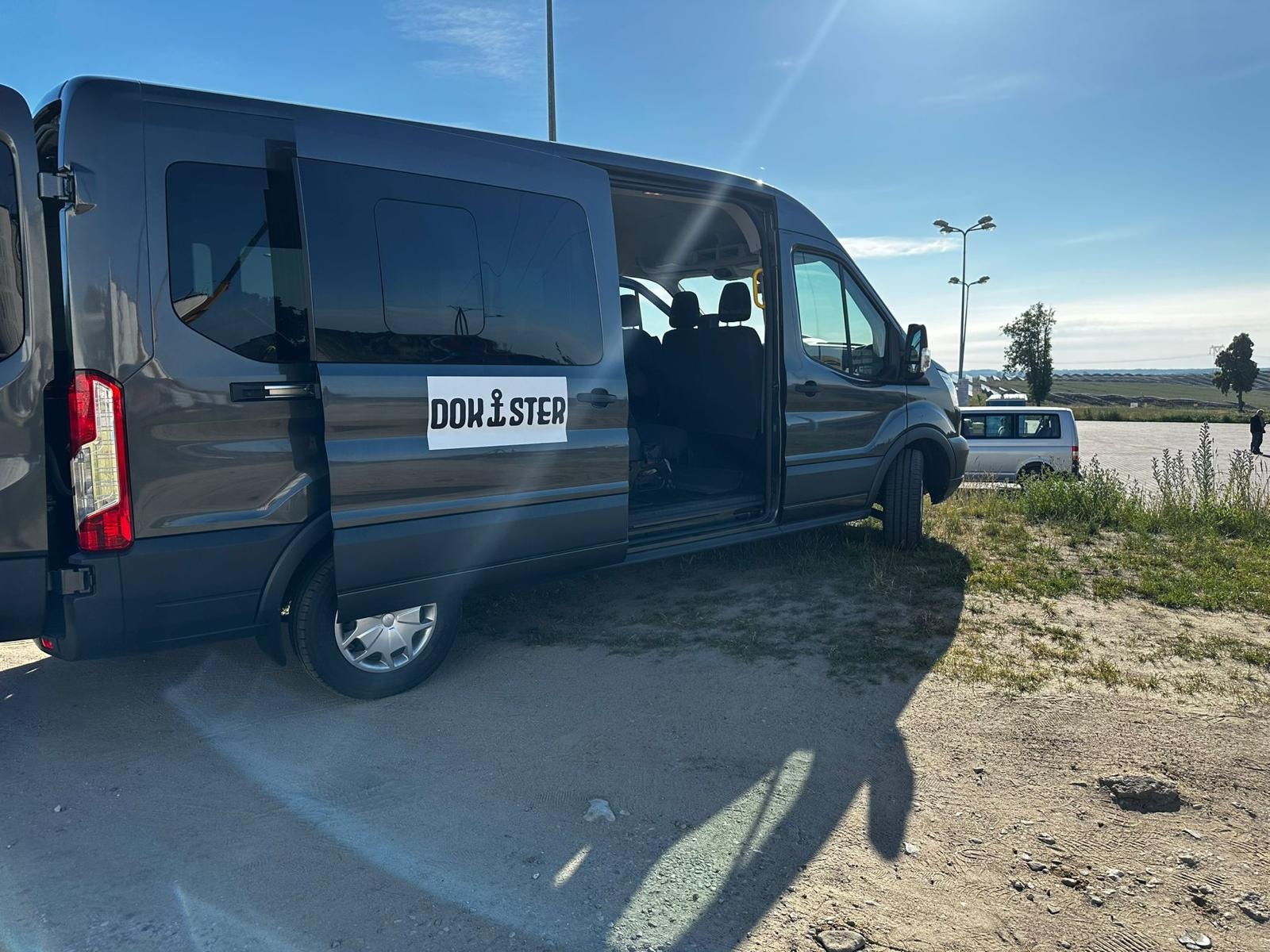 Szary van z otwartymi drzwiami bocznymi, logo firmy 'DOK STER' na boku, zaparkowany na parkingu w słoneczny dzień. Widoczne inne pojazdy i elementy krajobrazu.
