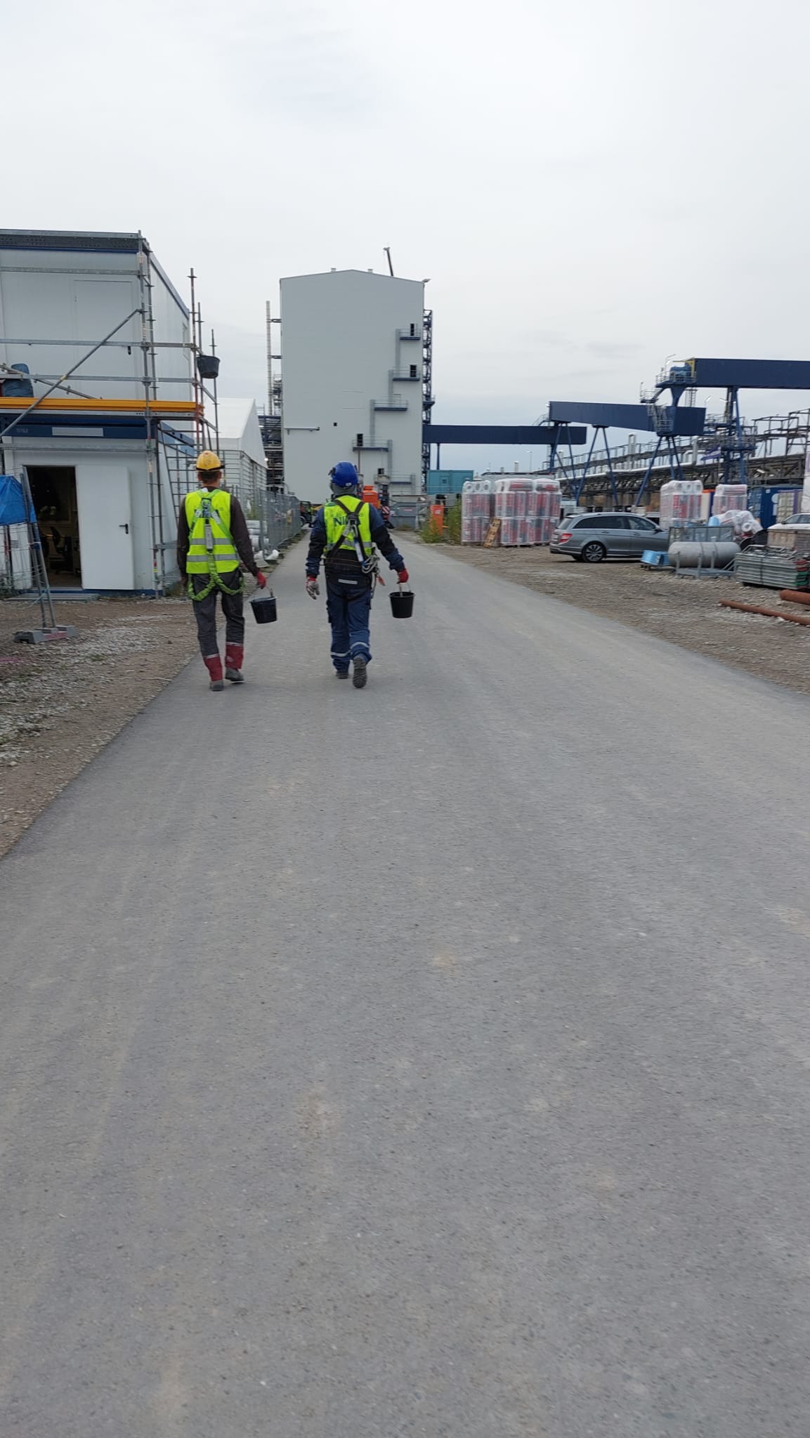 Dwóch pracowników w kamizelkach idzie drogą na tle konstrukcji budowlanej i materiałów. Pracownicy niosą wiadra. Widok z tyłu, pochmurne niebo.