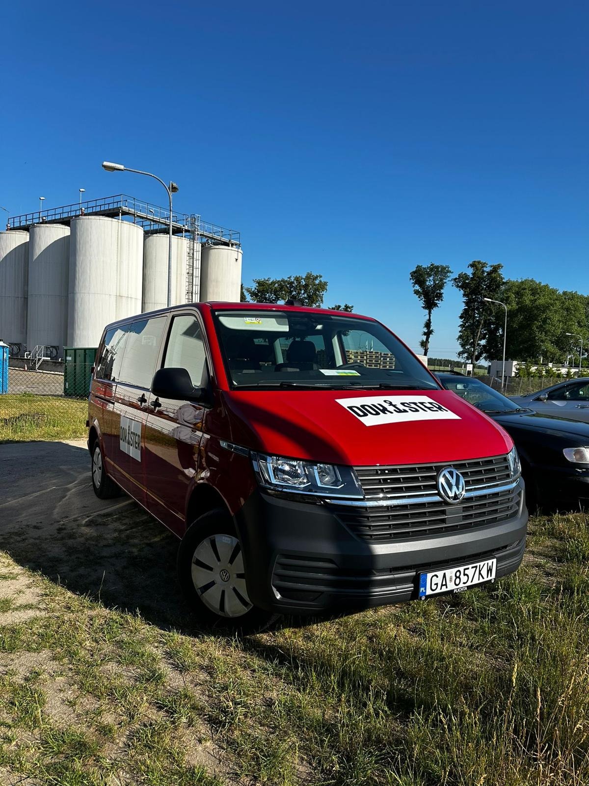 Czerwony bus firmy z naklejkami na tle silosów i błękitnego nieba. Pojazd z logotypem firmy, prawdopodobnie świadczącej usługi outsourcingu.