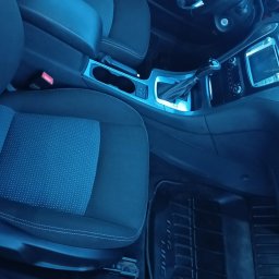 AutoGlanz - Autokosmetyka Konserwacja antykorozyjna Wulkanizacja - Wnętrze samochodu z widocznymi fotelami z tapicerką, deską rozdzielczą i dywanikami. Ciemna kolorystyka.