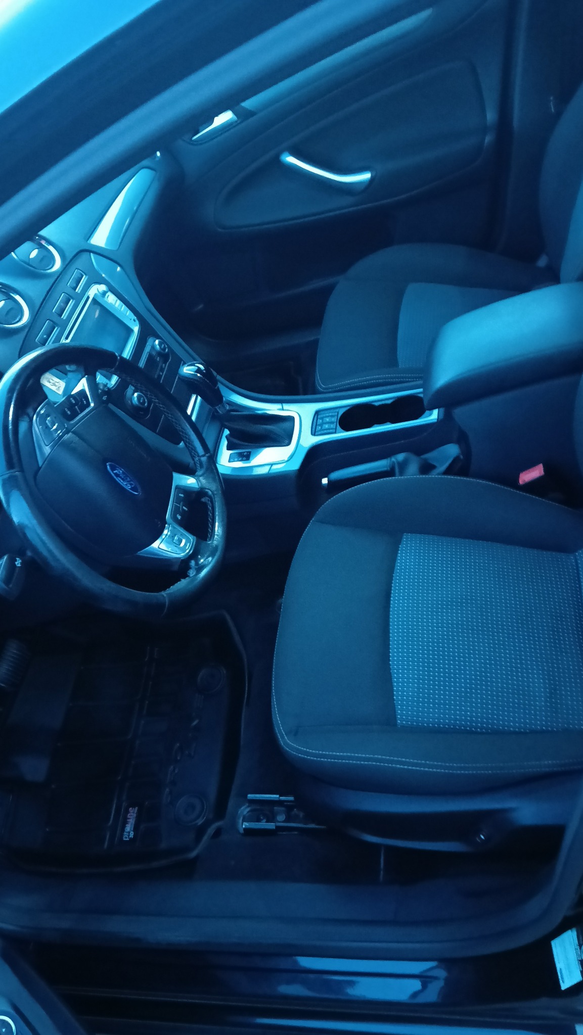 Wnętrze samochodu marki Ford z widocznymi fotelami pokrytymi ciemną tapicerką w drobny wzór, kierownicą z logo Forda i elementami deski rozdzielczej.