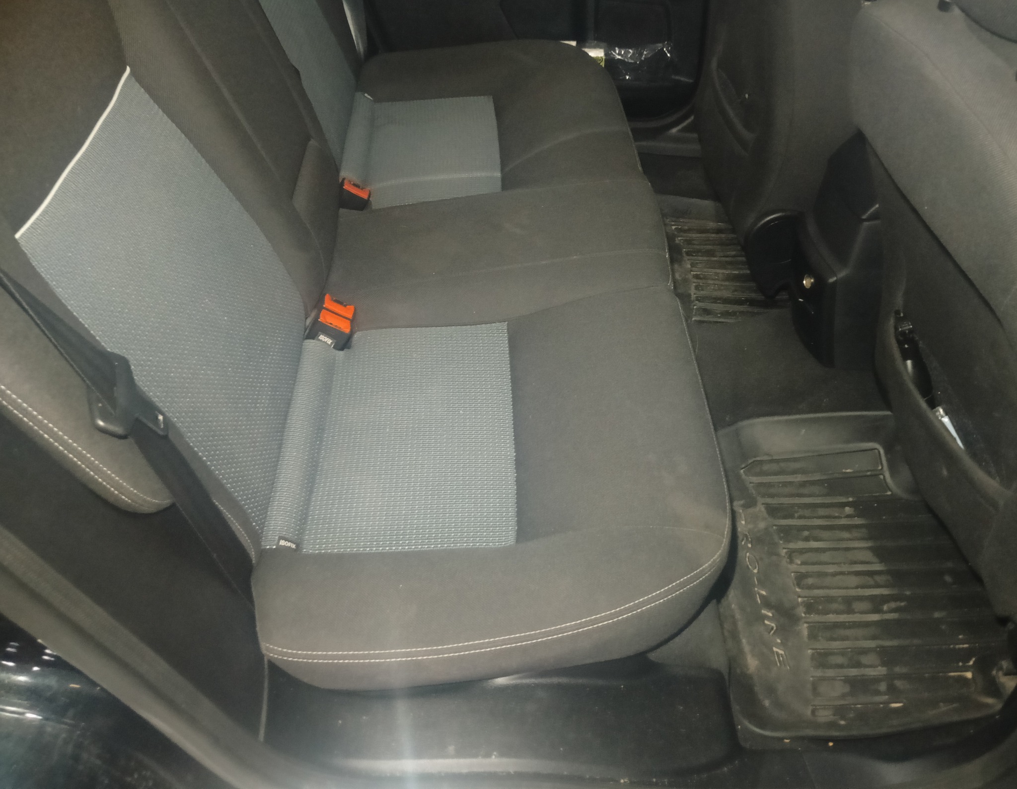 Ciemne, materiałowe siedzenia tylne samochodu z szarą wstawką i pomarańczowymi klamrami pasów. Widoczne gumowe dywaniki z napisem.