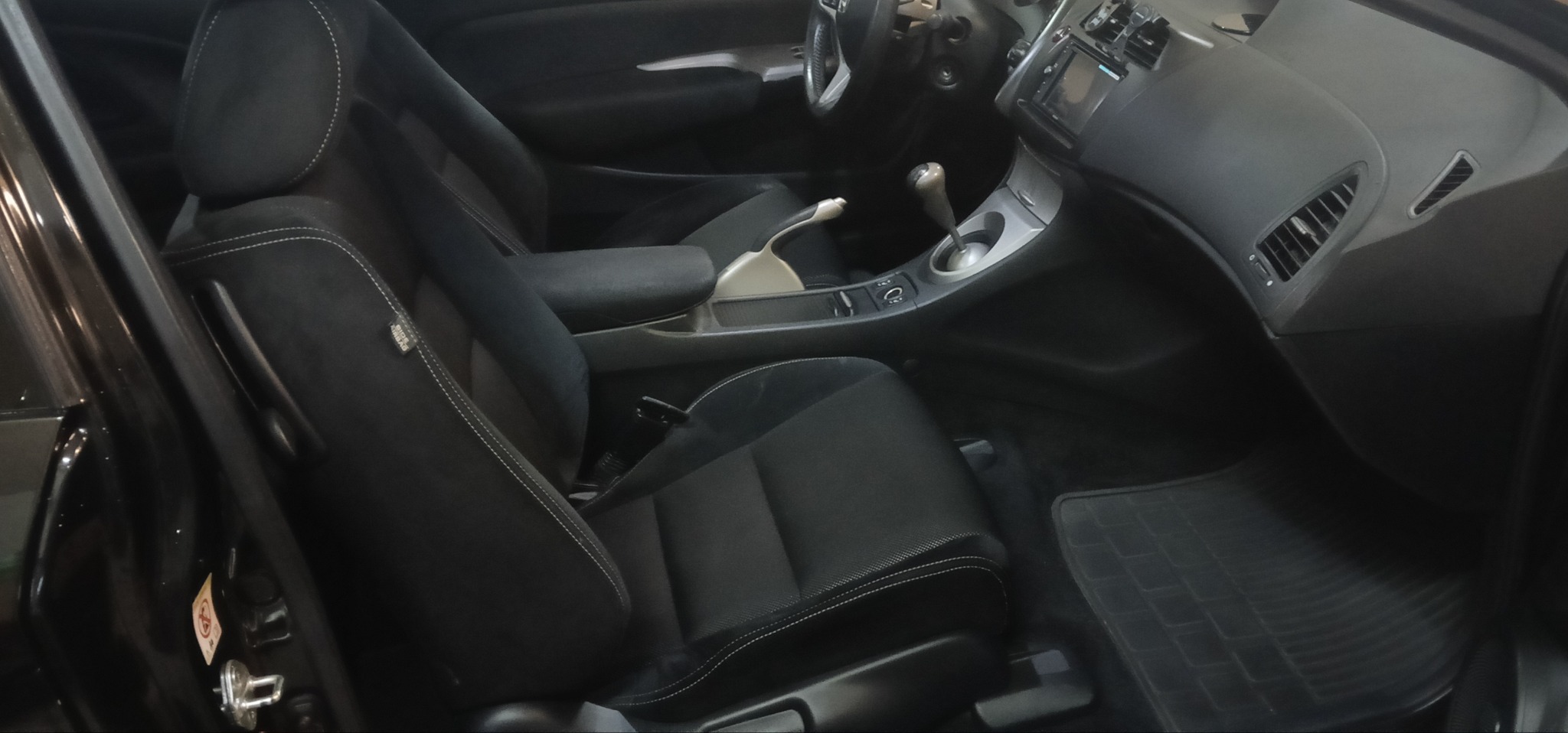 Wnętrze czarnego samochodu z wypranymi, czystymi fotelami z czarnej tapicerki z jasnymi przeszyciami. Widoczna deska rozdzielcza i dywaniki.