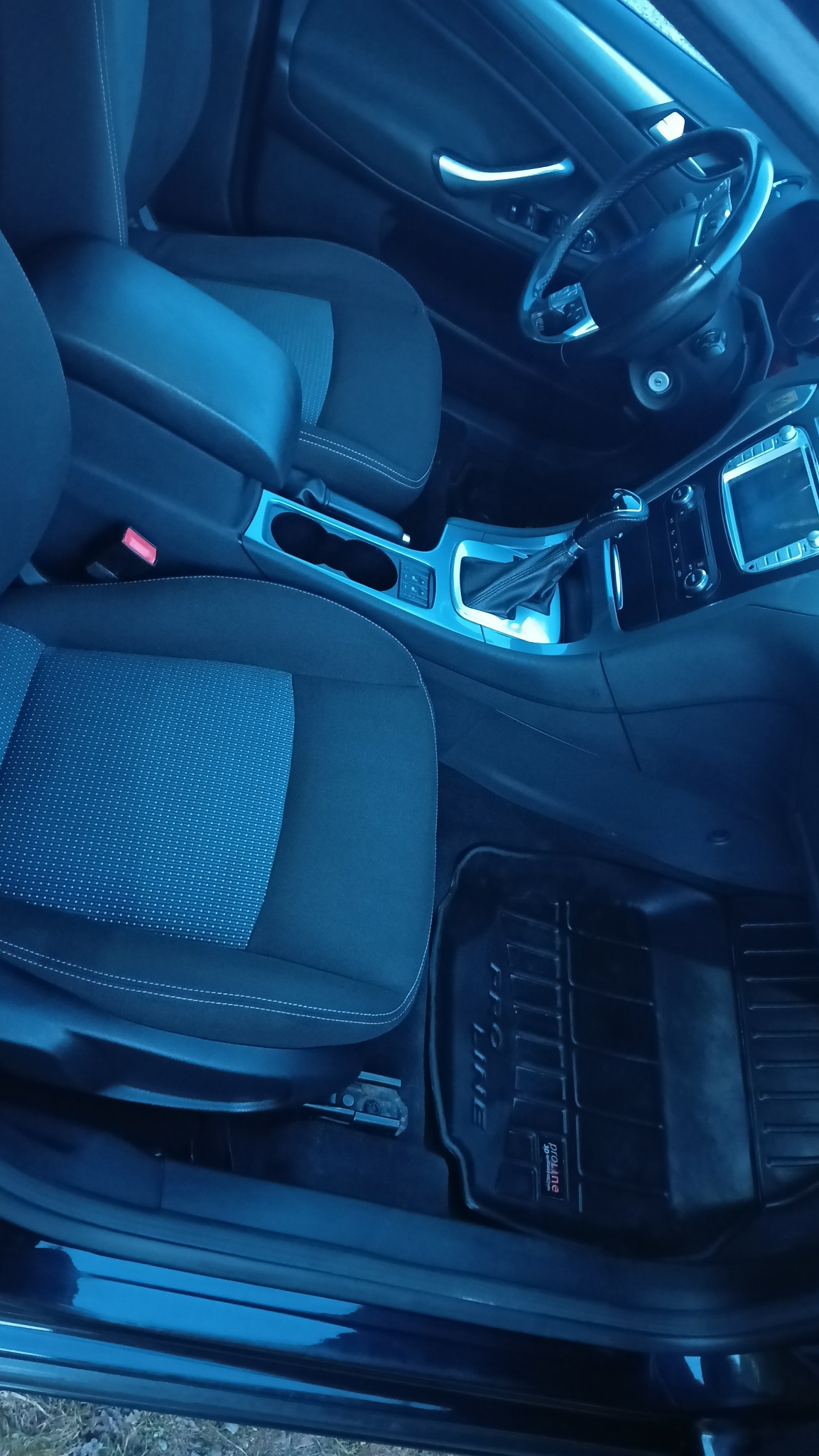 Wnętrze samochodu z widocznymi fotelami z tapicerką, deską rozdzielczą i dywanikami. Ciemna kolorystyka.