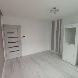 Witold Pawelec - Jasne wnętrze po remoncie: drewniana podłoga, białe meble, drzwi z przeszkleniami, ściana z fioletowymi pasami. Widok na sufit z lampą.