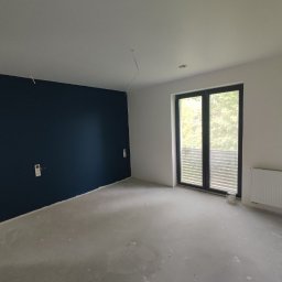 Witold Pawelec - Pokój po malowaniu ścian na biało i granatowo, z betonową podłogą i grzejnikiem. Widoczne gniazdka elektryczne i duże okno balkonowe.