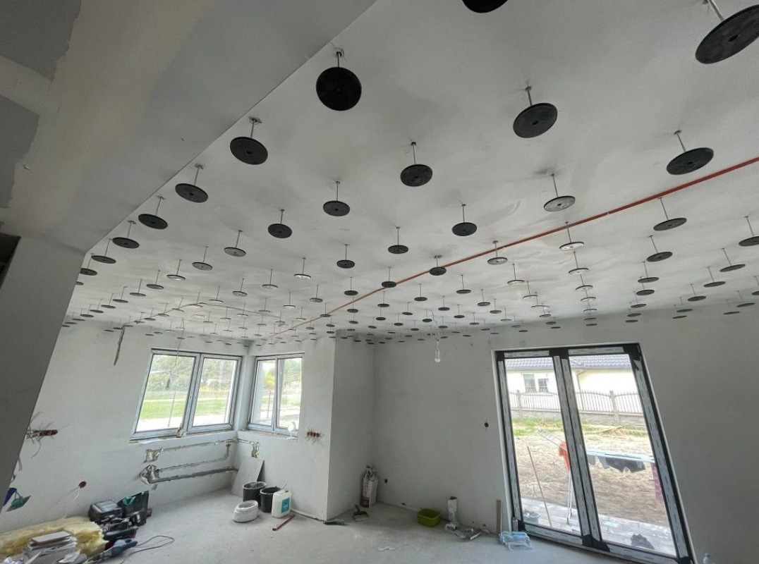 Sufit z zamocowanymi elementami izolacji akustycznej, przygotowany do montażu płyt. Widoczne okna i elementy instalacji elektrycznej.