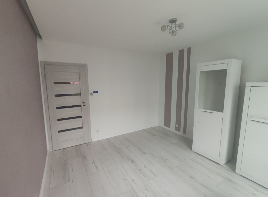 Jasne wnętrze po remoncie: drewniana podłoga, białe meble, drzwi z przeszkleniami, ściana z fioletowymi pasami. Widok na sufit z lampą.