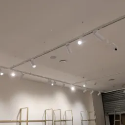 Białe reflektory szynowe LED zamontowane na suficie w sklepie. Widoczne elementy konstrukcyjne i oświetleniowe, w tle regały i zamknięta roleta.