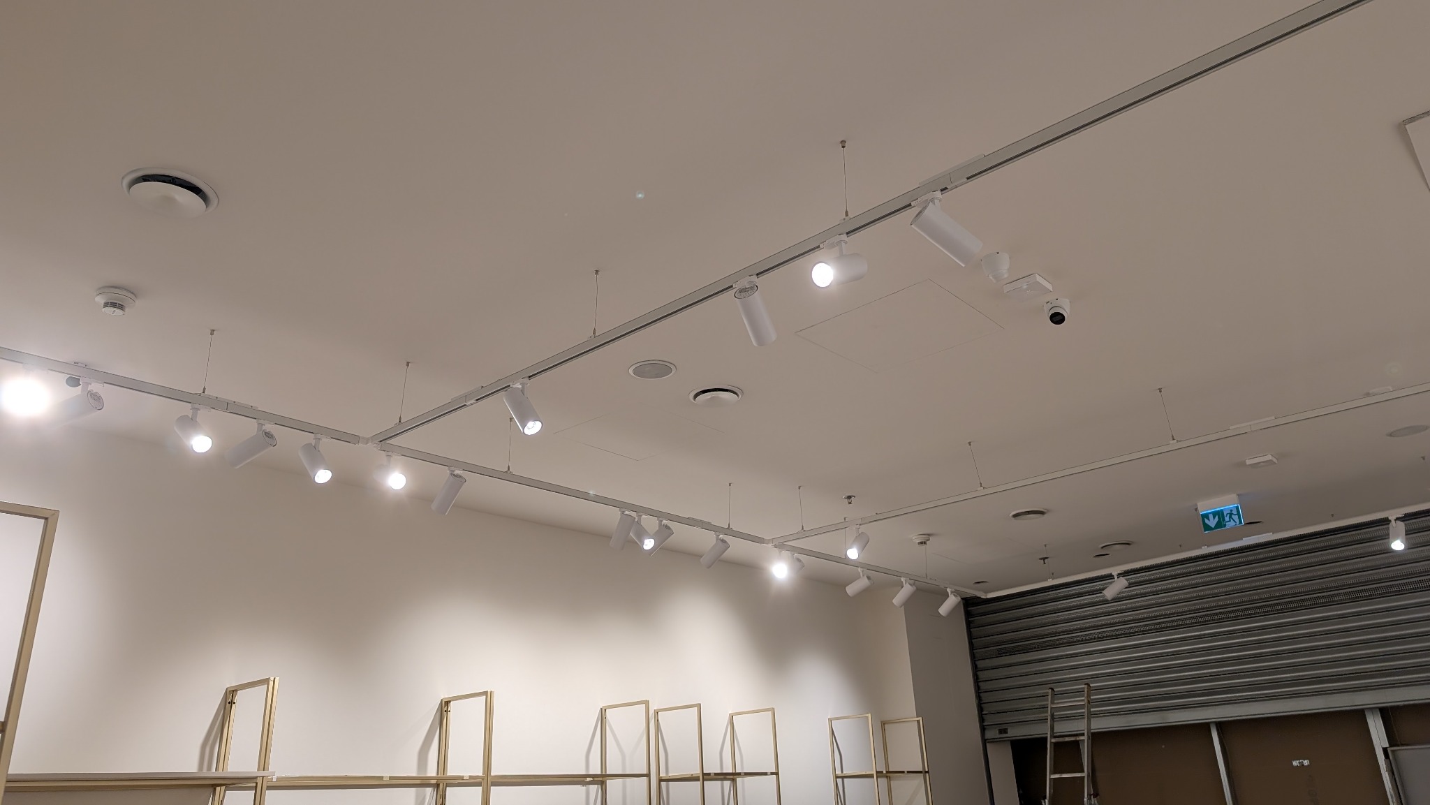 Białe reflektory szynowe LED zamontowane na suficie w sklepie. Widoczne elementy konstrukcyjne i oświetleniowe, w tle regały i zamknięta roleta.