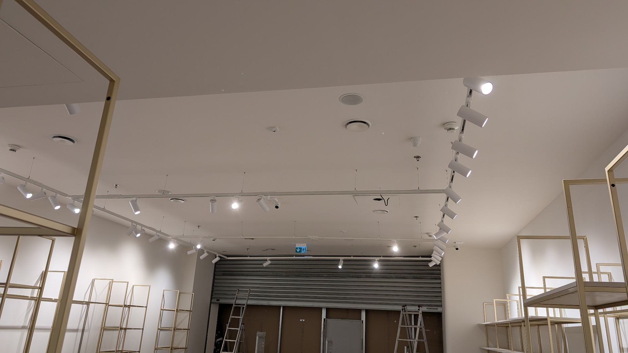 Białe reflektory szynowe zamontowane na suficie w sklepie z metalowymi regałami i zamkniętą roletą w tle, widok z dołu.