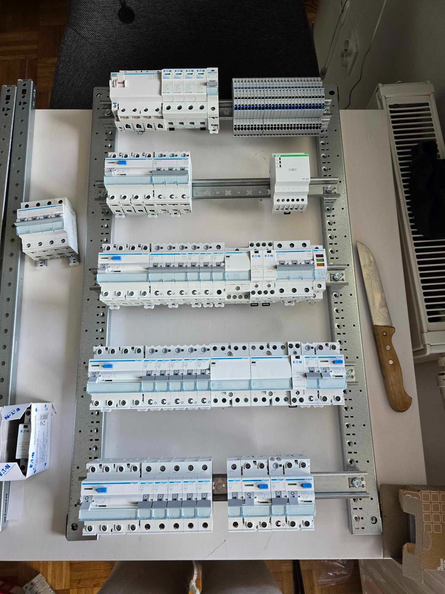 Rozdzielnica elektryczna z zamontowanymi bezpiecznikami Hager i EATON, widok z góry. Obok leży nóż z drewnianą rączką i kartonowe pudełko.