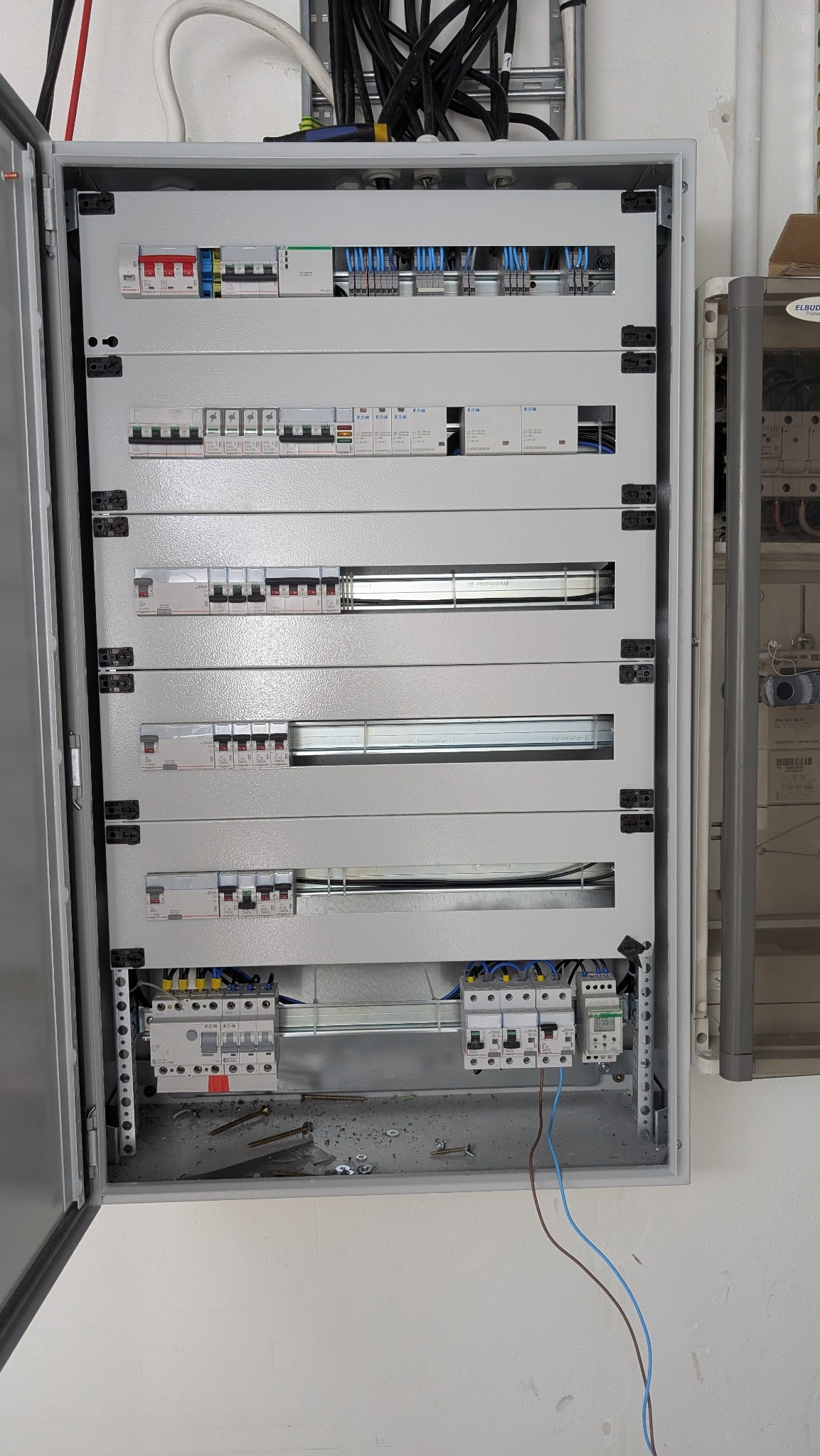 Otwarta szara szafa rozdzielcza z widocznymi bezpiecznikami, przewodami i elementami montażowymi. Widoczne przewody wychodzące z dołu szafy.